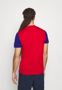 Man med långa dreadlocks som står vänd ifrån, iklädd en röd och blå kortärmad skjorta och mörka byxor mot en vit bakgrund.