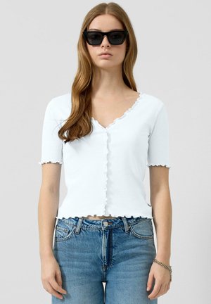 Młoda kobieta o długich włosach, nosząca czarne okulary przeciwsłoneczne w stylu cat-eye, białą, krótką bluzkę zapinaną na guziki oraz niebieskie dżinsy, na tle jednolitego tła.