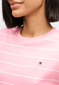 Ροζ κοντομάνικο πουκάμισο με οριζόντιες λευκές ρίγες, στρογγυλή λαιμόκοψη και μικρό λογότυπο με κόκκινα, λευκά και μπλε χρώματα στο στήθος.