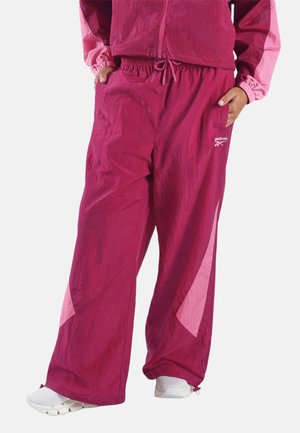 Model draagt roze en magenta wijde trainingsbroeken met elastische taille en zakken, gecombineerd met witte sportschoenen.