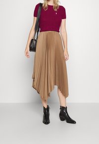 Mauve kortärmad tröja med strukturerat mönster, kombinerad med en kamelfärgad plisserad midikjol med hög-låg fåll. Svarta ankelboots fullbordar looken.