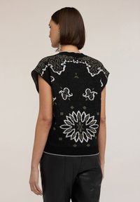 Suéter negro de punto con mangas cortas, intrincados patrones en blanco y oliva, y un dobladillo acanalado. La parte posterior presenta un diseño floral.