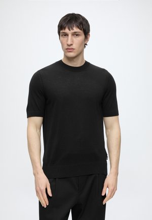 Joven con cabello corto y oscuro, vestido con una camiseta de punto negra y lisa de manga corta, posando frente a un fondo gris claro.