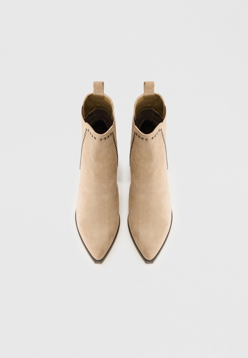 Beżowe zamszowe botki za kostkę z ostrymi noskami, ozdobnymi ćwiekami wzdłuż szwów oraz pętlami z tyłu ułatwiającymi zakładanie.