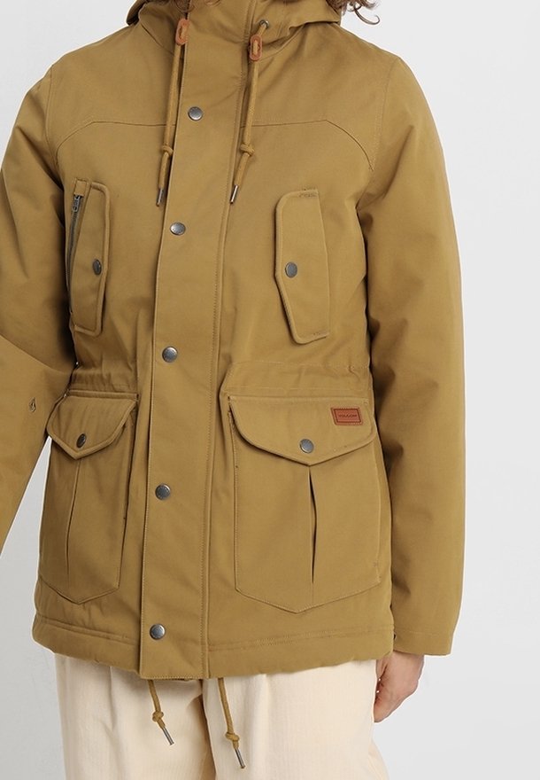 Veste à capuche couleur beige avec quatre poches à rabat à l'avant, fermetures par boutons, et cordons de serrage à la taille et à l'ourlet, portée avec un pantalon clair.