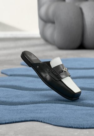 Mocassin à enfiler noir et blanc avec détail métallique reposant sur un tapis bleu texturé, avec un fauteuil gris flou en arrière-plan.