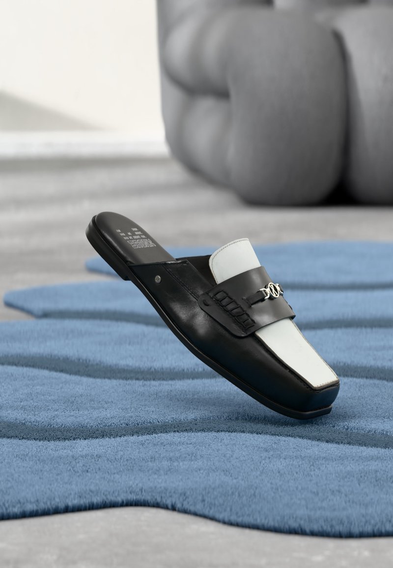 Mocassin à enfiler noir et blanc avec détail métallique reposant sur un tapis bleu texturé, avec un fauteuil gris flou en arrière-plan.