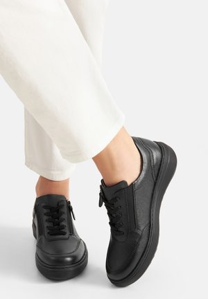 Czarne skórzane trampki z frontem na sznurowadła, bocznym zamkiem błyskawicznym, wyściełanym kołnierzem i teksturowaną powierzchnią, zestawione z białymi zwężanymi spodniami.