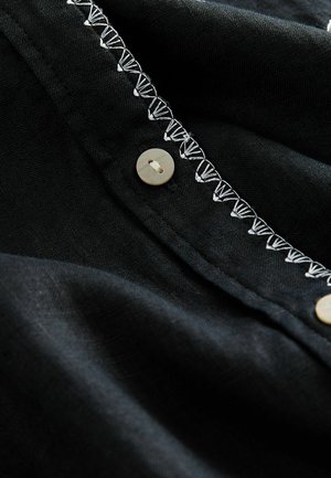 Sort stof i nærbillede med en knap og hvid dekorativ syning langs kanten, der fremhæver tekstur og designdetaljer.