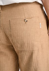 Ljusbruna byxor med herringbone-textur, försedda med en bakficka som stängs med en brun knapp och en liten tygetikett på sidan.