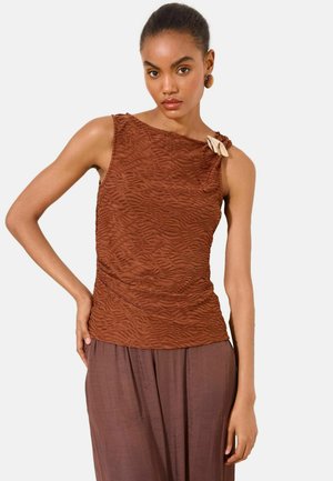 Femme portant un haut marron texturé sans manches avec un nœud sur l'épaule gauche et une jupe marron ample, posant avec la main sur la hanche.