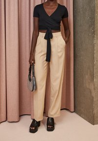 Černý zavinovací top s krátkými rukávy a zavazováním v pase, doplněný béžovými širokými kalhotami. Doplňky zahrnují černou kabelku s cvoky a boty s masivním podpatkem.