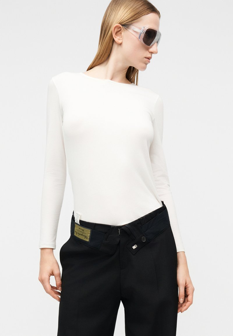 Femme portant des lunettes de soleil oversize transparentes, un haut blanc ajusté à manches longues et un pantalon noir ample avec une taille non boutonnée.