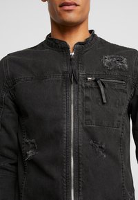 Mies, jolla on päällään musta kulunut farkkutakki, jossa on etuvetoketju, vetoketjullinen rintatasku ja rispaantuneet tikkaukset rinnassa ja alhaalla.