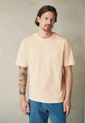 Bărbat cu păr de lungime medie și barbă poartă o cămașă pală de culoarea piersicii cu mâneci scurte și buzunar, arătând tatuaje cu natură și fulg de nea pe antebrațul stâng.