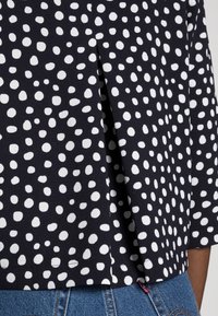 Černá látková blůza s bílými nepravidelnými puntíky, která má mírně volný střih a detail bočního rozparku na lemu pro větší pohyb.
