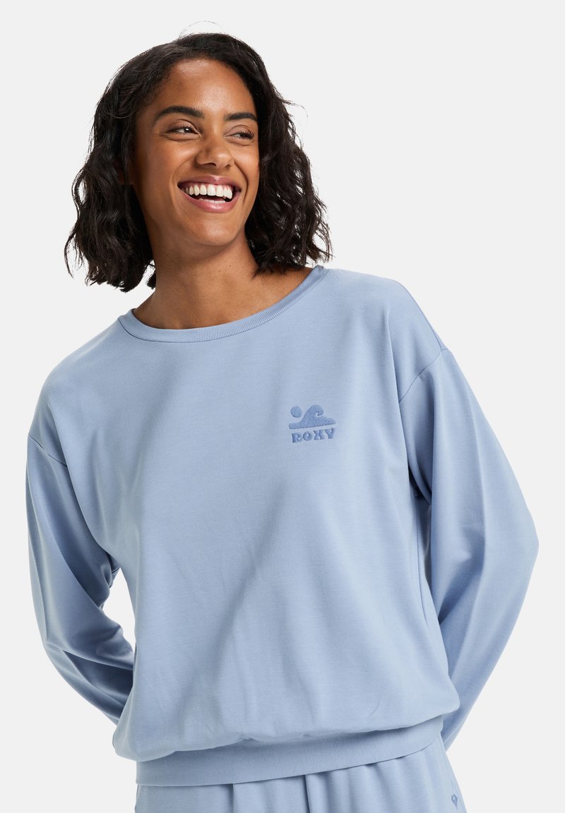 Světle modrá mikina vyrobená z měkké látky. Má kulatý výstřih, volný střih a malé vyšívané logo na přední straně.