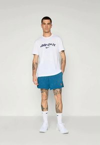 Λευκό βαμβακερό T-shirt με ναυτικό λογότυπο "Just Do It", συνδυασμένο με μπλε σορτς και λευκά αθλητικά παπούτσια. Το μοντέλο έχει ορατά τατουάζ στα χέρια.