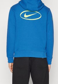 Modrá mikina s kapucí vyrobená z bavlněné směsi. Na zadní straně má velké žluté logo Nike a manžety a spodní lem jsou žebrované.