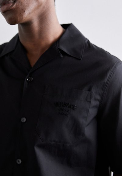 Chemise noire à manches longues avec un col ajusté, ornée d'un logo brodé sur la poche poitrine et d'une texture lisse et mate.