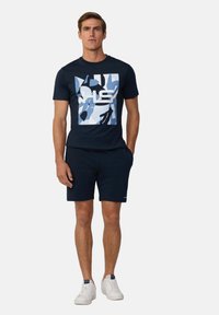 Navy katoenen T-shirt met abstract blauw en witte grafiek, gecombineerd met marineblauwe shorts en witte sneakers. Casual, fitted ontwerp.
