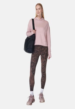 Jonge vrouw die een roze hoodie draagt, bruine leggings met slangprint, roze sneakers, witte sokken en een zwarte schoudertas.