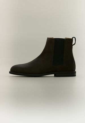 Dunkelbrauner Veloursleder-Chelsea-Stiefel mit schwarzem elastischem Seitenteil und Zuglasche auf einer flachen schwarzen Sohle vor neutralem Hintergrund.