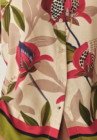 Beige stof met grote rode, groene en bruine bloem- en bladrprints, met crèmekleurige knopen en een rode en groene rand nabij de zoom.