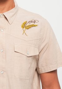 Beige short-sleeve shirt with two front pockets, snap buttons, and embroidered yellow wheat design on the left chest. Text reads "Tough."