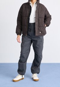 Veste matelassée brun à carreaux avec col montant, associée à un pantalon cargo ample gris foncé et des baskets blanches avec des semelles beiges.