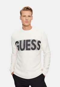Maglione bianco a maglia con un grande logo "GUESS" in paillettes nere sul petto. Polsini e orlo a coste con un design a girocollo.