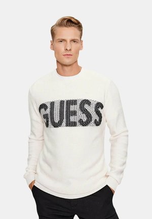 Maglione bianco a maglia con un grande logo "GUESS" in paillettes nere sul petto. Polsini e orlo a coste con un design a girocollo.