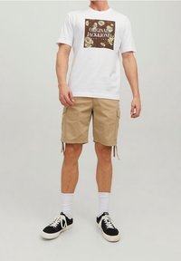 Valkoinen t-paita, jossa on ruskea kukkakuvio, beige cargo-shortsit ja mustat lenkkarit, joissa on valkoisia yksityiskohtia, tyyliteltynä valkoisilla sukilla.