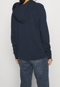 Felpa blu navy in misto cotone con tasca a marsupio e cappuccio con cordino regolabile; abbinata a jeans in denim azzurro.