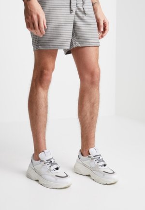 Baskets blanches de sport avec des accents gris, design à lacets et semelle texturée, associées à un short à carreaux noir et blanc.