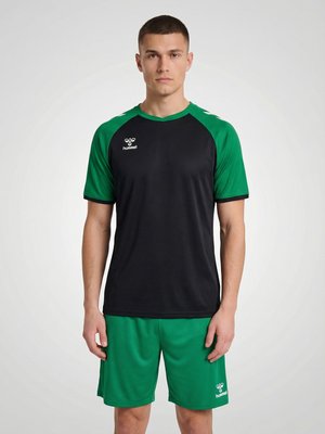 Ung mand stående fremadvendt iført sort og grøn hummel sportstrøje og grønne shorts mod ensfarvet lyst baggrund.