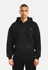 Uomo che indossa una felpa nera con cerniera e tasche frontali, in piedi contro uno sfondo chiaro e uniforme, con uno sguardo neutro.