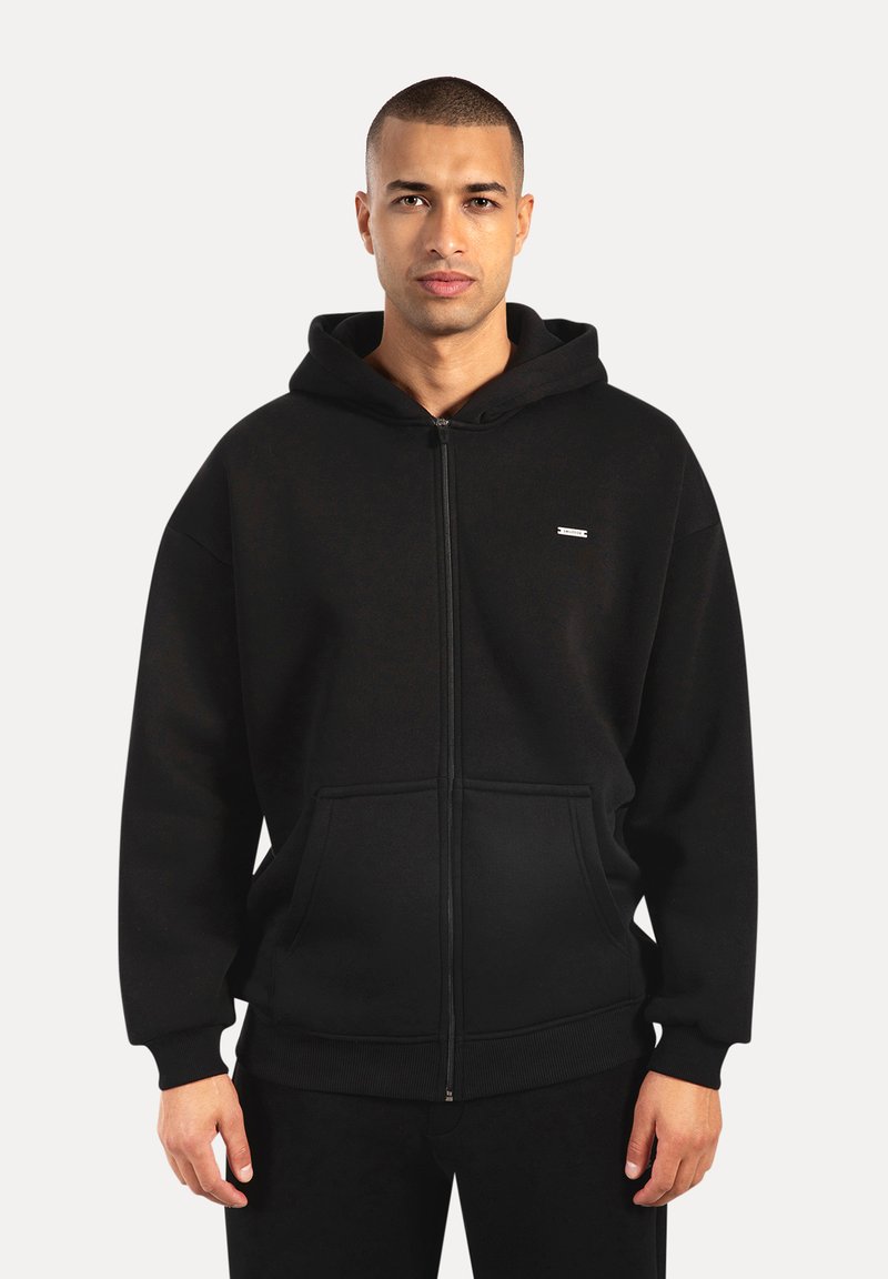 Uomo che indossa una felpa nera con cerniera e tasche frontali, in piedi contro uno sfondo chiaro e uniforme, con uno sguardo neutro.