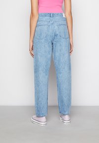 Jeans de ganga azul claro com uma cintura alta, apresentando um corte reto, dois bolsos traseiros e um acabamento desgastado. Usados com sapatilhas cor-de-rosa.