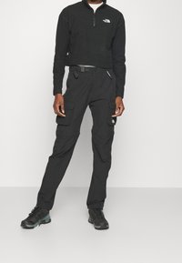 Schwarzes Fleece-Top mit Viertelreißverschluss und Logo, kombiniert mit schwarzen Cargo-Hosen mit Taschen und einem Gürtel, ergänzt durch schwarze Wanderschuhe.