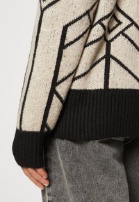 Stickad tröja med en krämfärgad botten och svart geometrisk design, med en svart ribbad nederkant. Mjuk textur med kontrasterande mönster.