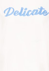 T-shirt en tissu blanc avec texte brodé en bleu indiquant "Délicat." Le design présente une texture lisse et une typographie décontractée.