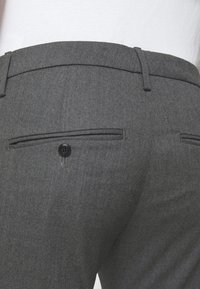 Vue de dos d'une personne portant un pantalon de costume gris foncé avec des poches passepoilées boutonnées et des passants de ceinture, associé à une chemise blanche.