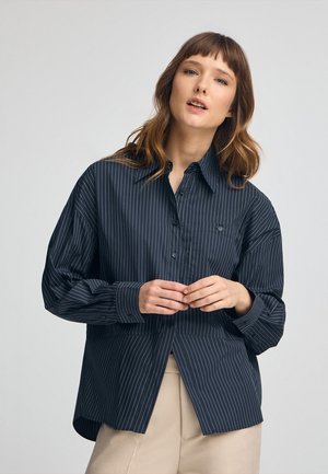 Camicia abbottonata blu navy con strisce verticali bianche, caratterizzata da un colletto classico, una vestibilità rilassata e una tasca singola sul petto. Texture del tessuto morbida.