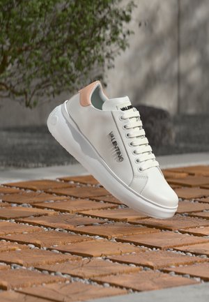 Baskets en cuir blanc avec un accent rose clair, dotées d'un bout arrondi, de lacets plats et d'un logo sur le côté, posées sur une surface en bois.