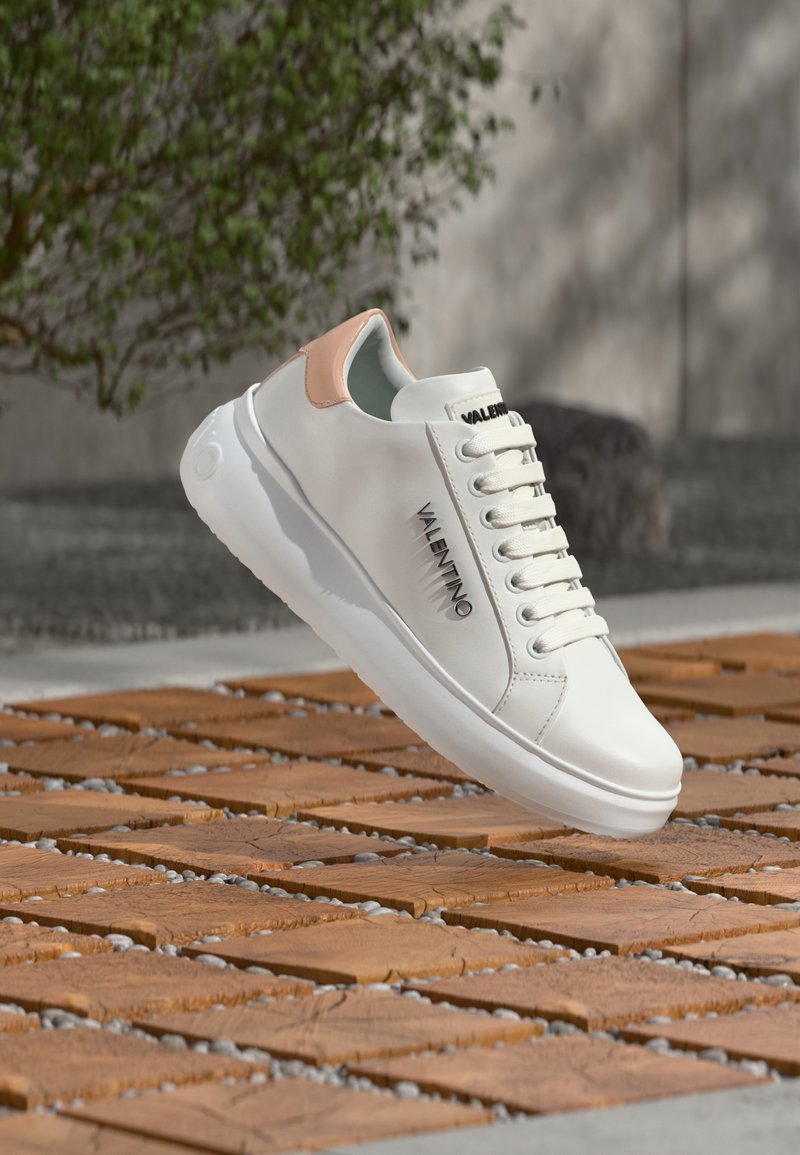 Baskets en cuir blanc avec un accent rose clair, dotées d'un bout arrondi, de lacets plats et d'un logo sur le côté, posées sur une surface en bois.