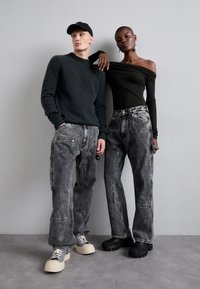 Grå striktrøje, off-shoulder sort top, lysegrå og mørkegrå baggy jeans, chunky sneakers og strukturerede sorte sko. Minimalistisk baggrund.