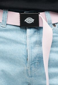 Φωτεινά γαλάζια τζιν με υφασμάτινη ζώνη σε παστέλ ροζ χρώμα, με μαύρη μεταλλική αγκράφα που φέρει ανάγλυφη την επιγραφή "Dickies." Κοντινή λήψη της περιοχής της μέσης.