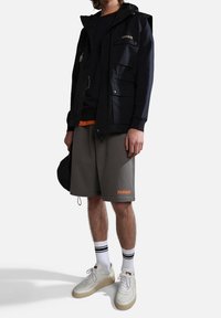 Chaqueta negra de nailon con múltiples bolsillos, pantalones cortos grises, zapatillas blancas y calcetines a rayas; presenta detalles en naranja en los pantalones cortos y en la chaqueta.
