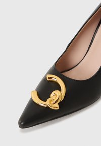 Soulier noir en cuir à talons hauts, avec un bout pointu et un accent en métal doré sur le cou-de-pied, présentant une texture lisse et un design élégant.